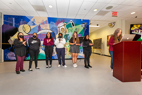 Adjunct Professor and accomplished mural artist Julia Cocuzza ‘11 M.F.A., who taught the experimental class that will officially be offered Fall 2023, addresses the crowd with students involved with creating the mural (left to right) Elizabeth Li, Katherine Infante, Crystal Gonzalez, Sasecie Bernard, Angela Auriti, and Nicole Harripersad.